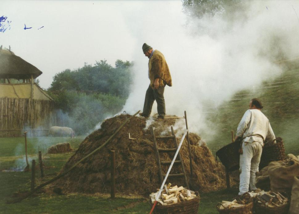 11. 2de fase: Na een uur wordt de deksel eraf gehaald. Met de lange porstok wordt alle verbrande hout naar beneden gepord. Er worden weer 2 manden nieuw aanmaakhout in gegooid.