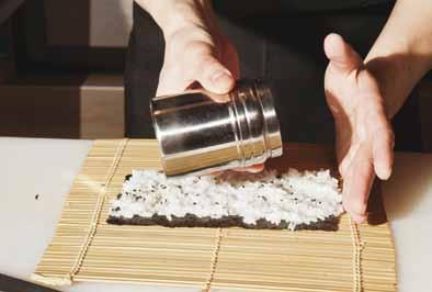 Fabrieksreportage 报导 De nori wordt op een rieten matje gelegd, wat straks nodig is voor het oprollen. Vanuit de rijst-pan wordt een klein beetje rijst gepakt.
