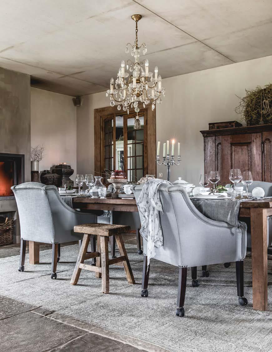 Boven de eettafel hangt een brocante kroonluchter uit Frankrijk, de tafel en stoelen zijn antiek. Het kleed is van Hoffz. De houtkachel op de achtergrond verwarmt het hele huis.