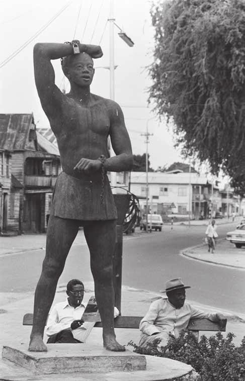 Inzet: mannen lezen in 1975 de krant bij het standbeeld Kwakoe in Paramaribo dat de afschaffing van de slavernij op 1 juli 1863 herdenkt (Nationaal Archief).