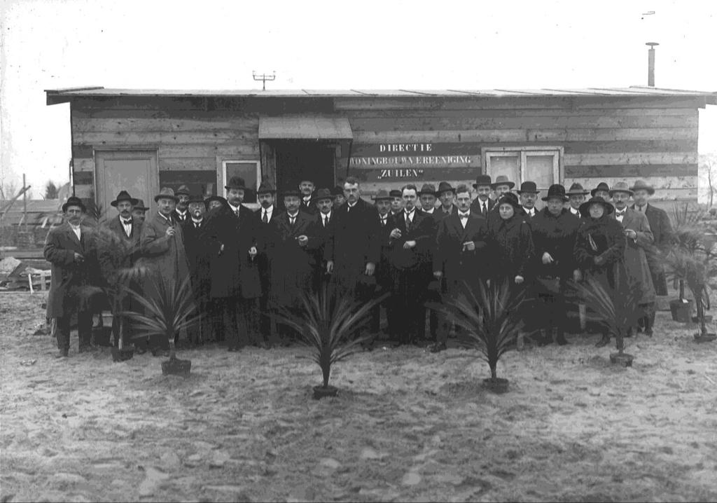 De medewerkers van woningbouwvereniging Zuilen stellen zich aan u voor, staande voor de directiekeet, achter een rij wuivende palmen.