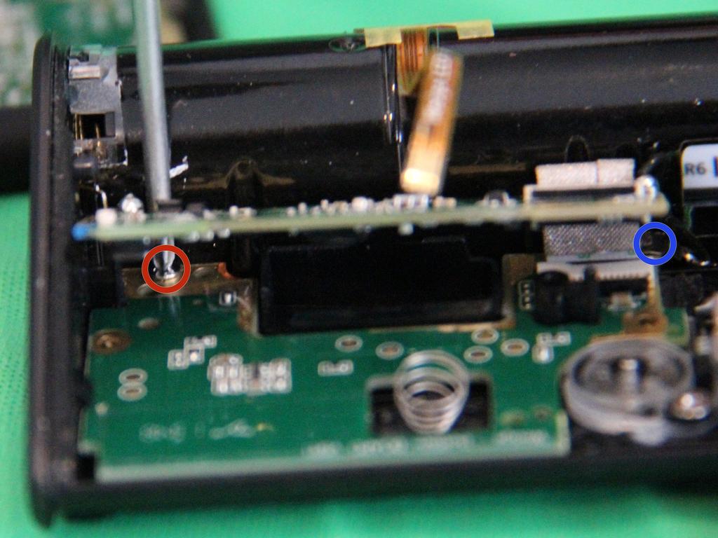 Schuif een kleine phillips schroevendraaier tussen de bovenste printplaat en het batterijcompartiment om de schroef rood omcirkeld te verwijderen.
